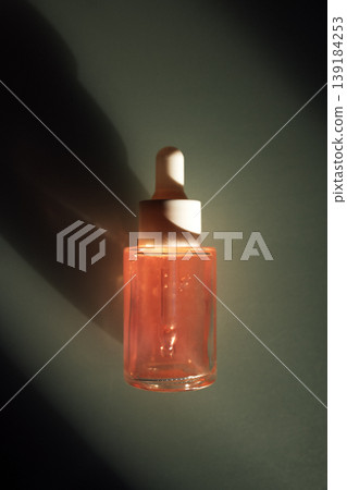 Glass dropper bottle filled with pink serum, featuring a white dropper cap, illuminated by soft light against a dark green background Glass dropper bottle filled with pink serum, featuring a white dropper cap, illuminated by soft light against a dark green background 139184253