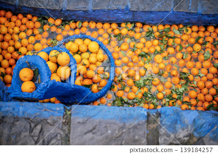 Fresh oranges in a blue basket surrounded by scattered oranges in a stone basin, with green leaves visible in the water Fresh oranges in a blue basket surrounded by scattered oranges in a stone basin, with green leaves visible in the water 139184257
