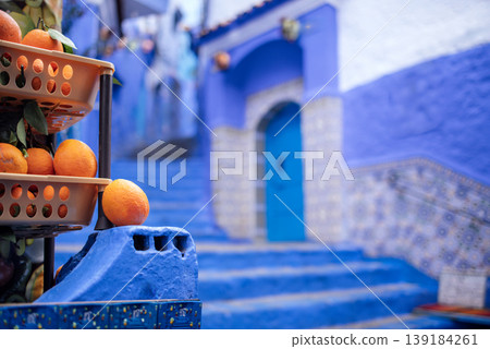 Orange fruits in a basket on blue stone steps leading to a blue door in a vibrant blue and white alleyway of Chefchaouen, Morocco 139184261