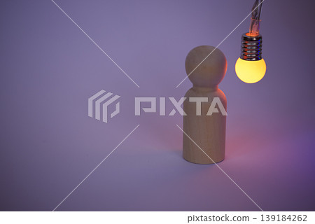 Wooden figurine standing on a plain surface illuminated by a hanging light bulb with a warm yellow glow against a soft purple background Wooden figurine standing on a plain surface illuminated by a hanging light bulb with a warm yellow glow against a soft purple background 139184262