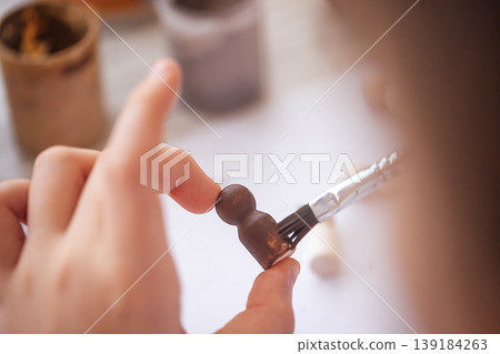 Close-up of a hand applying paint to a small wooden figurine with a brush, surrounded by jars of paint in a creative workspace setting Close-up of a hand applying paint to a small wooden figurine with a brush, surrounded by jars of paint in a creative workspace setting 139184263