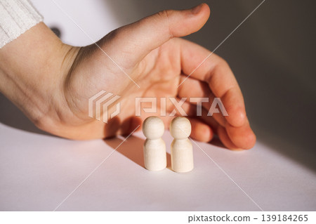 Hand reaching towards two wooden figurines on a white surface, casting soft shadows, with a blurred background creating a minimalistic aesthetic 139184265