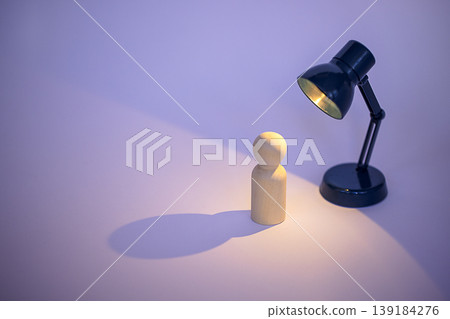 Wooden figurine stands under a black desk lamp casting a warm light and shadow on a smooth surface with a soft purple background Wooden figurine stands under a black desk lamp casting a warm light and shadow on a smooth surface with a soft purple background 139184276