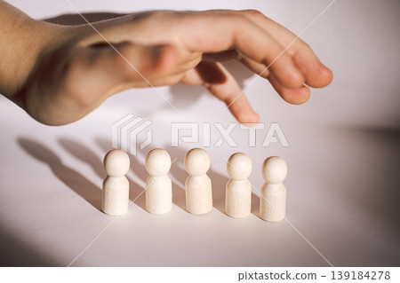 Hand reaching over five wooden figurines arranged in a row on a light surface, casting shadows, emphasizing the interaction between the hand and figures Hand reaching over five wooden figurines arranged in a row on a light surface, casting shadows, emphasizing the interaction between the hand and figures 139184278