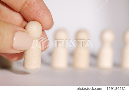 Hand holding a wooden figurine in front of a row of similar wooden figures on a light background, showcasing a minimalist design and simple shapes 139184281