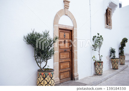 Wooden door framed by white wall, adorned with decorative planters, showcasing vibrant greenery and intricate patterns, creating a charming and inviting atmosphere in a serene outdoor setting Wooden door framed by white wall, adorned with decorative planters, showcasing vibrant greenery and intricate patterns, creating a charming and inviting atmosphere in a serene outdoor setting 139184330