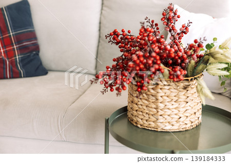 Decorative arrangement of vibrant red berries and greenery in a woven basket on a stylish green table beside a cozy couch, enhancing interior decor aesthetics Decorative arrangement of vibrant red berries and greenery in a woven basket on a stylish green table beside a cozy couch, enhancing interior decor aesthetics 139184333
