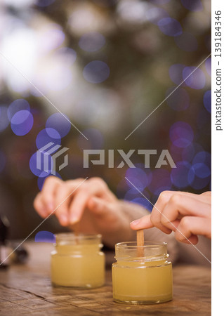 Hands of a person stirring yellow candle wax in glass jars on a wooden table, with a blurred background of colorful bokeh lights creating a warm, inviting atmosphere Hands of a person stirring yellow candle wax in glass jars on a wooden table, with a blurred background of colorful bokeh lights creating a warm, inviting atmosphere 139184346