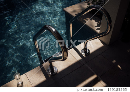 Stainless steel pool handrails beside clear blue water with sunlight reflections on the surface and a glass of water on the edge of the pool area Stainless steel pool handrails beside clear blue water with sunlight reflections on the surface and a glass of water on the edge of the pool area 139185265