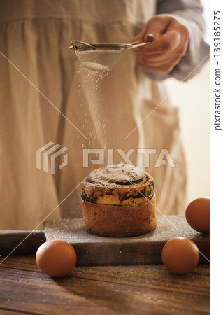 Person in beige apron sifting powdered sugar over freshly baked cake on wooden table with eggs and rustic kitchen background Person in beige apron sifting powdered sugar over freshly baked cake on wooden table with eggs and rustic kitchen background 139185275