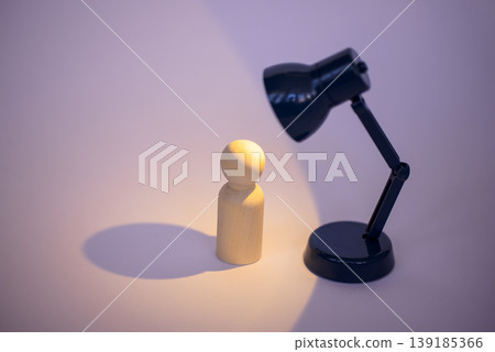 Wooden figurine illuminated by a black desk lamp casting a soft shadow on a light purple background, creating a minimalist and artistic composition 139185366