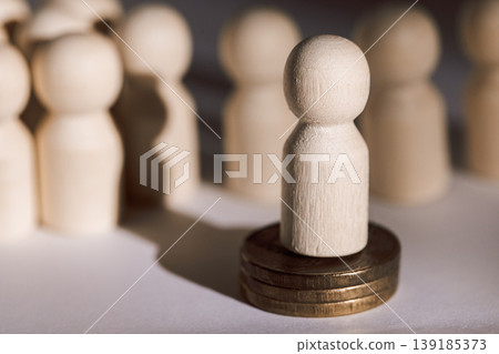 Wooden figurine standing on coins surrounded by blurred wooden figures, representing individuality and financial concepts in a minimalist setting Wooden figurine standing on coins surrounded by blurred wooden figures, representing individuality and financial concepts in a minimalist setting 139185373