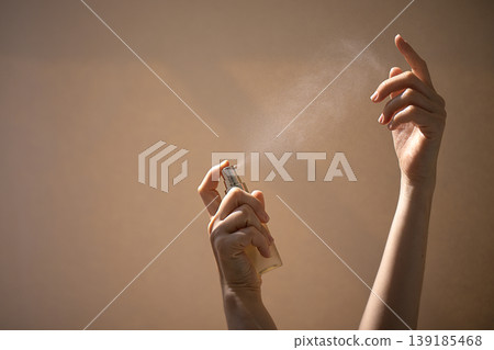 Female hands holding a spray bottle releasing mist against a soft, neutral background, showcasing beauty or skincare product application in a serene setting Female hands holding a spray bottle releasing mist against a soft, neutral background, showcasing beauty or skincare product application in a serene setting 139185468
