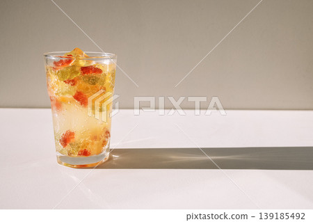 Close-up of a glass of mineral water with delicious marmalade bears and ice cubes on a white table. A cup of cool, bright, delicious sweet drink on a light background. Close-up of a glass of mineral water with delicious marmalade bears and ice cubes on a white table. A cup of cool, bright, delicious sweet drink on a light background. 139185492