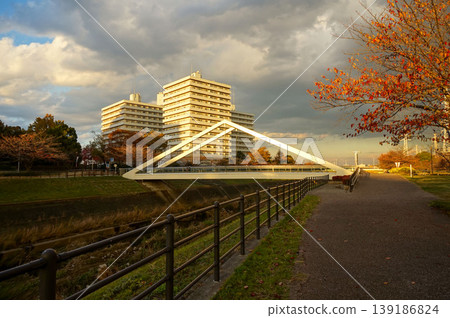 A Japanese cityscape with a bridge and river illuminated by the autumn evening light, and striking autumn leaves and clouds. 139186824