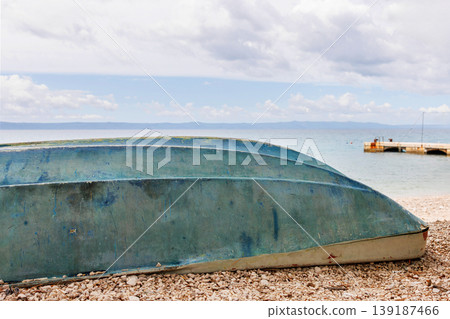 Overturned blue boat rests on a pebble beach beside calm sea water. Adriatic sea coast in Makarska, Croatia. Minimal composition with copy space. Overturned blue boat rests on a pebble beach beside calm sea water. Adriatic sea coast in Makarska, Croatia. Minimal composition with copy space. 139187466