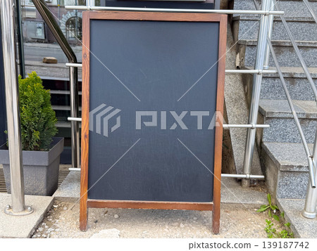 Blank chalkboard sign standing beside stairs near a building entrance. Copy space, cafe menu, street advertising, small business frontage, and urban communication. Blank chalkboard sign standing beside stairs near a building entrance. Copy space, cafe menu, street advertising, small business frontage, and urban communication. 139187742