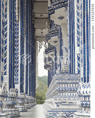 A grand corridor of sapphire and ivory pillars unfolds at Wat Paknam Khaem Nu, the 'Blue Temple.' These towering, mosaic-clad columns create a rhythmic perspective that draws the eye toward the lush 139187839