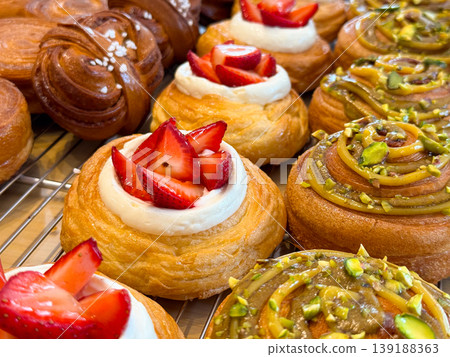 Danish pastries with strawberry cream and pistachio topping arranged closely on bakery rack. Sweet dessert selection, pastry variety, confectionery display and food styling concept. Danish pastries with strawberry cream and pistachio topping arranged closely on bakery rack. Sweet dessert selection, pastry variety, confectionery display and food styling concept. 139188363