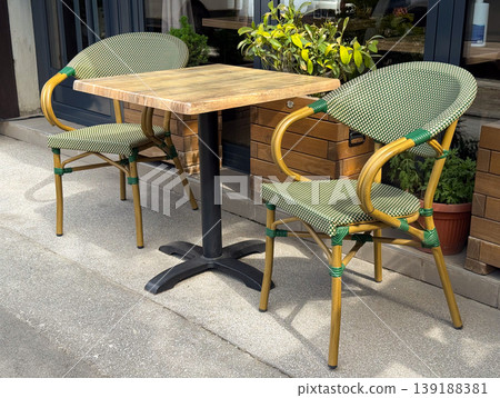 Empty cafe table with two woven bistro chairs on a sunny sidewalk terrace. Outdoor dining, hospitality setting, urban leisure and small business street furniture. Empty cafe table with two woven bistro chairs on a sunny sidewalk terrace. Outdoor dining, hospitality setting, urban leisure and small business street furniture. 139188381