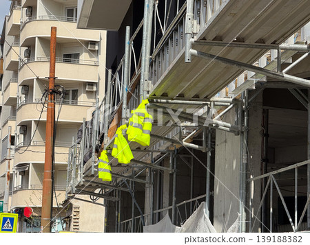 Construction scaffolding with fluorescent safety vests hanging near an unfinished building facade. Urban renovation, worker protection, building repair and construction site infrastructure. Construction scaffolding with fluorescent safety vests hanging near an unfinished building facade. Urban renovation, worker protection, building repair and construction site infrastructure. 139188382