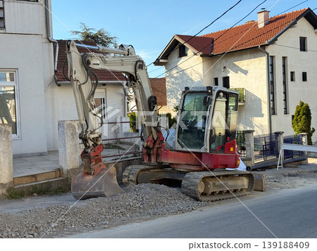 Compact excavator parked beside houses during road repair. Urban maintenance, residential construction, infrastructure renewal, machinery work, utility access, and neighborhood disruption. 139188409
