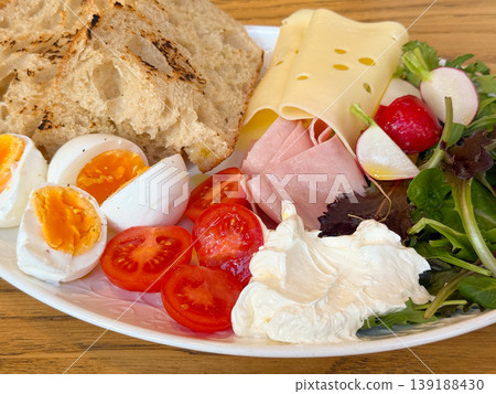 Breakfast plate with emmental cheese, ham, egg, tomato, radish, salad, cream and grilled bread on a wooden table. Morning meal, cafe dining, protein, vegetable variety and brunch nutrition. Breakfast plate with emmental cheese, ham, egg, tomato, radish, salad, cream and grilled bread on a wooden table. Morning meal, cafe dining, protein, vegetable variety and brunch nutrition. 139188430