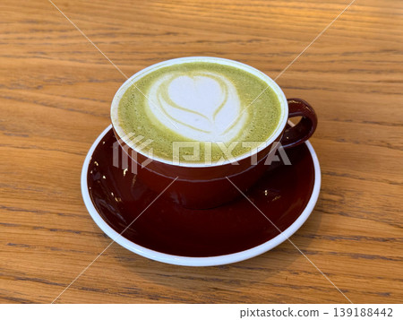 Matcha latte with heart foam art served in red cup on wooden table. Cafe beverage culture, barista craft, coffee alternative, and minimal composition. Matcha latte with heart foam art served in red cup on wooden table. Cafe beverage culture, barista craft, coffee alternative, and minimal composition. 139188442