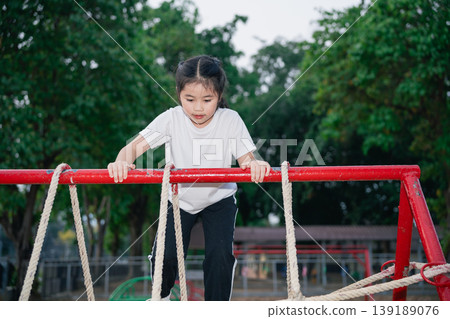 Young Girl Enjoying Outdoor Play While Climbing on Playground Equipment in a Sunny Park Surrounded by Green Trees and Nature's Beauty 139189076