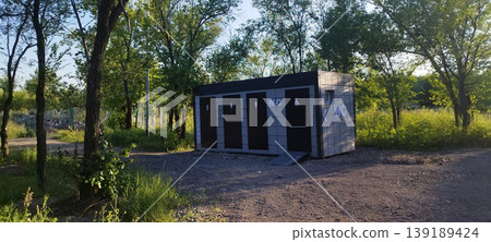 Modern rectangular public restroom is located in sunlit wooded area. Modular gray black structure features four separate doors with white "WC" signs, accessibility icons, including Modern rectangular public restroom is located in sunlit wooded area. Modular gray black structure features four separate doors with white "WC" signs, accessibility icons, including 139189424