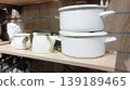 Set of minimalist white enamel cookware displayed on a wooden shelf. The collection includes stacked pots with lids and a small saucepan with a wooden handle, perfect for a modern, clean kitchen 139189465