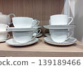Elegant white ceramic teacups and saucers stacked neatly on a light wooden shelf. The minimalist design features a thin dark rim, creating a sophisticated and clean look for kitchen or cafe decor 139189466