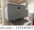 Close-up shows a grey herringbone fabric storage box with a metal handle, displayed on a retail shelf. It provides a stylish, practical solution for home organization and wardrobe management 139189467
