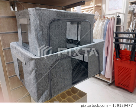 Two grey fabric storage bins with clear windows and zippers are stacked on a shelf in a store. They feature a herringbone pattern, offering an organized and stylish solution for home wardrobe storage 139189477