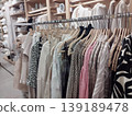 Neatly organized clothing rack in a modern retail store. A variety of patterned and neutral-toned garments hang on wooden hangers, reflecting a cozy, stylish aesthetic 139189478