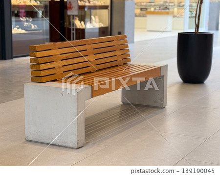 Empty wooden bench with concrete supports in a shopping mall corridor with blurred people and kiosks behind. Public furniture, waiting, urban commerce, rest and indoor transit 139190045