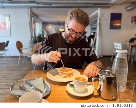 Smiling man with beard and glasses holding fork with dessert at cafe table with tea and tableware. Casual dining, enjoyment, social interaction, leisure moment and food experience. Smiling man with beard and glasses holding fork with dessert at cafe table with tea and tableware. Casual dining, enjoyment, social interaction, leisure moment and food experience. 139190318