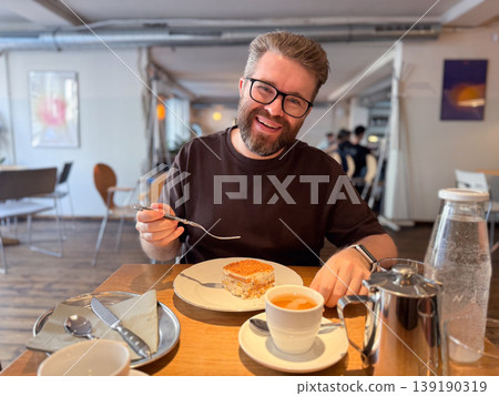 Smiling man with beard and glasses holding fork with dessert at cafe table with tea and tableware. Casual dining, enjoyment, social interaction, leisure moment and food experience. Smiling man with beard and glasses holding fork with dessert at cafe table with tea and tableware. Casual dining, enjoyment, social interaction, leisure moment and food experience. 139190319