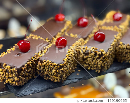 Chocolate cake slices with cherry topping and nut coating in display. Dessert indulgence, pastry craftsmanship, sweet decoration, confectionery assortment, and retail presentation. Chocolate cake slices with cherry topping and nut coating in display. Dessert indulgence, pastry craftsmanship, sweet decoration, confectionery assortment, and retail presentation. 139190665