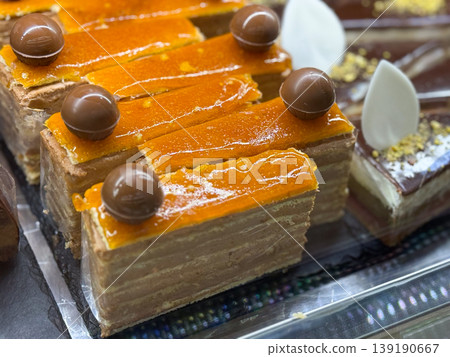 Layered caramel glazed cake squares topped with chocolate spheres in bakery display. Dessert craftsmanship, confectionery texture, sweet indulgence, pastry layering, and retail presentation of gourmet Layered caramel glazed cake squares topped with chocolate spheres in bakery display. Dessert craftsmanship, confectionery texture, sweet indulgence, pastry layering, and retail presentation of gourmet 139190667
