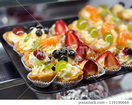 Assorted fruit tartlets with cream and berries in bakery display close-up. Dessert variety, pastry assortment, fresh ingredients, confectionery selection, and retail food presentation concept. Assorted fruit tartlets with cream and berries in bakery display close-up. Dessert variety, pastry assortment, fresh ingredients, confectionery selection, and retail food presentation concept. 139190668