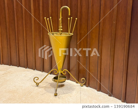 Vintage brass umbrella stand placed near wooden wall paneling. Interior decor, storage object, metal craftsmanship, hallway accessory, functional design, and classic furniture element in indoor 139190684