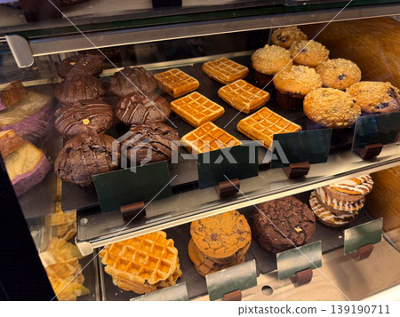 Assorted pastries and waffles displayed inside bakery showcase. Food selection, dessert variety, sweet snack, retail presentation, cafe interior, and consumer choice concept. 139190711