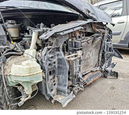 A car front end is damaged after a traffic accident. The metal is crumpled and the radiator is broken. Other cars are parked nearby on the roadside, indicating an urban area. 139191254