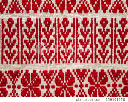 Close up view of white linen fabric featuring bright red geometric embroidery. The design shows classic patterns commonly found in Slavic or Eastern European art. Close up view of white linen fabric featuring bright red geometric embroidery. The design shows classic patterns commonly found in Slavic or Eastern European art. 139191258