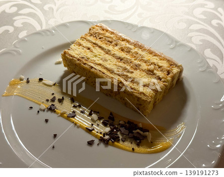 Layered cake slice with caramel sauce, chocolate flakes, and almond pieces on a white dessert plate. Pastry serving, sweet course, restaurant dessert, confectionery texture, and indulgent treat. Layered cake slice with caramel sauce, chocolate flakes, and almond pieces on a white dessert plate. Pastry serving, sweet course, restaurant dessert, confectionery texture, and indulgent treat. 139191273
