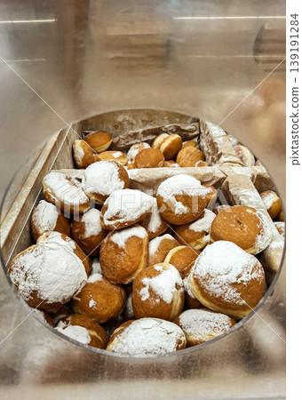 A glass container holds many traditional deep fried donuts covered in powdered sugar. The display is part of a pastry shop. The setting invites customers to enjoy sweet treats. A glass container holds many traditional deep fried donuts covered in powdered sugar. The display is part of a pastry shop. The setting invites customers to enjoy sweet treats. 139191284