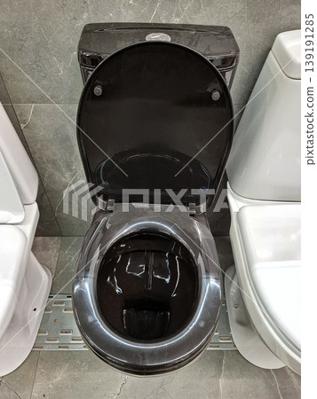 A black ceramic toilet is placed in a store. It features a simple design and glossy finish. The toilet is situated beside a white model on a grey marble tile backdrop. 139191285