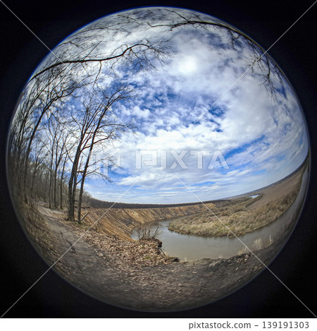 A winding river flows through a valley surrounded by bare trees. The sky is filled with clouds, showing the change of seasons in springtime. A winding river flows through a valley surrounded by bare trees. The sky is filled with clouds, showing the change of seasons in springtime. 139191303