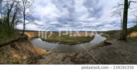 A wide view shows a winding river shaped like a horseshoe from a high cliff. The river flows through green fields under a cloudy sky in early spring. A wide view shows a winding river shaped like a horseshoe from a high cliff. The river flows through green fields under a cloudy sky in early spring. 139191350